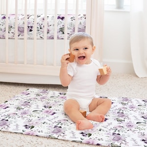 May include: A rectangular play mat featuring a white background with a repeating pattern of purple floral blossoms, green leaves, and the name Rosalie in black script. The mat is made of fabric and is laid flat on a carpeted floor.