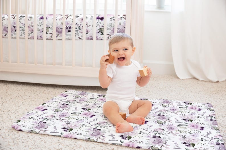 May include: A rectangular play mat featuring a white background with a repeating pattern of purple floral blossoms, green leaves, and the name Rosalie in black script. The mat is made of fabric and is laid flat on a carpeted floor.