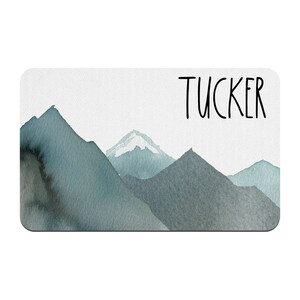 May include: A watercolor painting of a mountain range with a snowy peak. The word "TUCKER" is written in black at the top of the image.