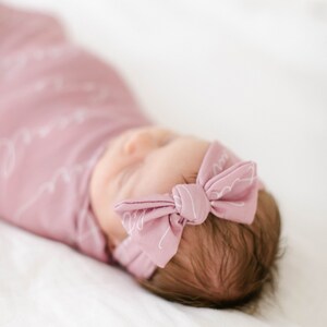 May include: A baby wrapped in a soft, light pink swaddle blanket with a matching bow headband. The swaddle and headband feature a delicate white script pattern. The baby is lying on a white surface.