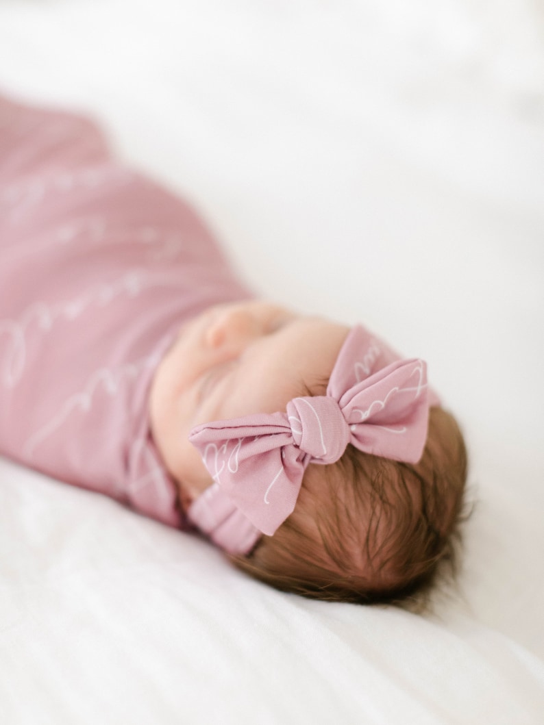 May include: A baby wrapped in a soft, light pink swaddle blanket with a matching bow headband. The swaddle and headband feature a delicate white script pattern. The baby is lying on a white surface.