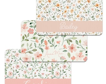 Pode incluir: Três tapetes de mesa personalizados com um design floral. Os tapetes de mesa têm uma borda rosa com um nome escrito em branco em itálico. Os nomes são Bailey, Anna Mae e Cookie.