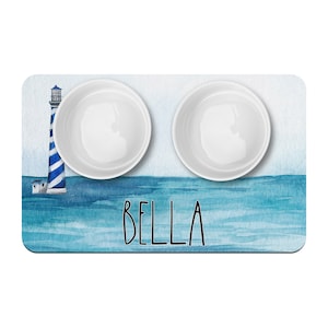 May include: A blue and white watercolor pet bowl mat with a lighthouse and the name "BELLA" printed on it. Two white ceramic bowls are on the mat.