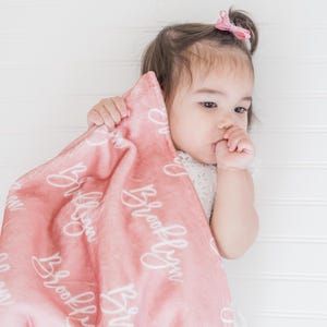 May include: A pink baby blanket with the name "Brooklyn" written in white cursive script. A baby girl is holding the blanket and looking to the side.