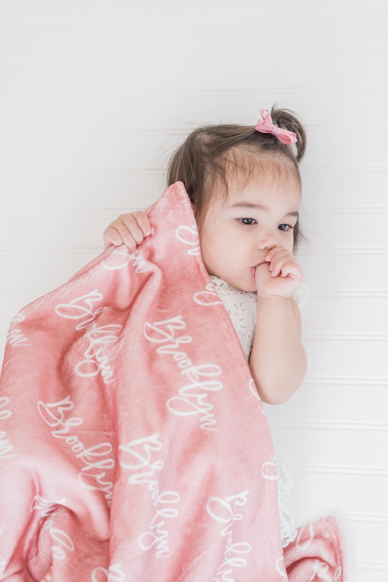 May include: A pink baby blanket with the name "Brooklyn" written in white cursive script. A baby girl is holding the blanket and looking to the side.