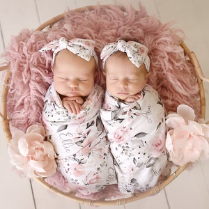 K&ouml;nnte beinhalten: Zwei Neugeborene, eingewickelt in florale Swaddles mit ihren Namen Ellie und Ava, die darauf gestickt sind. Sie tragen passende florale Stirnb&auml;nder und schlafen friedlich in einem Weidenkorb, der mit rosa Fell ausgelegt ist.