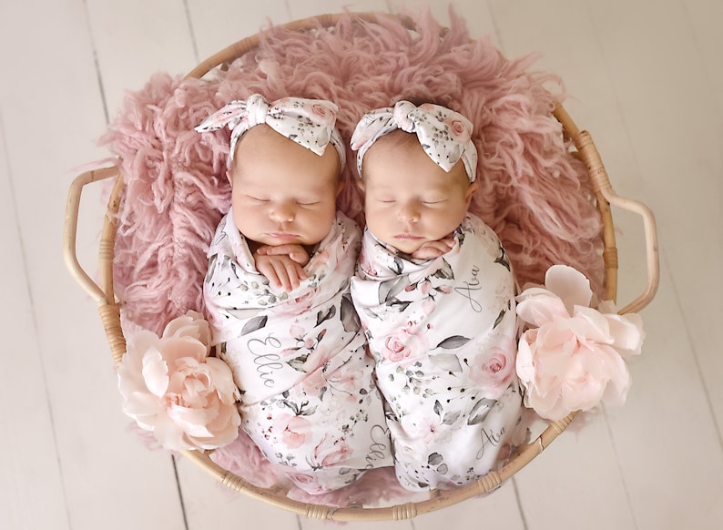 K&ouml;nnte beinhalten: Zwei Neugeborene, eingewickelt in florale Swaddles mit ihren Namen Ellie und Ava, die darauf gestickt sind. Sie tragen passende florale Stirnb&auml;nder und schlafen friedlich in einem Weidenkorb, der mit rosa Fell ausgelegt ist.