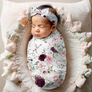 May include: A white swaddle blanket with a floral pattern in shades of pink and burgundy. The blanket is wrapped around a baby, and the baby is wearing a headband with a floral bow. The blanket has the name "Sophia" written on it in cursive.
