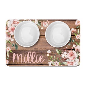 May include: A wooden pet food bowl mat with a floral design in shades of pink and green. The mat has two white ceramic bowls and the name "millie" written in pink.