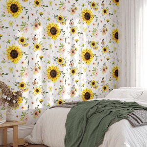 May include: Wallpaper featuring a repeating pattern of bright yellow sunflowers, soft pink flowers, and green foliage on a white background. The wallpaper is applied to a wall behind a bed with white bedding and a dark green throw blanket.