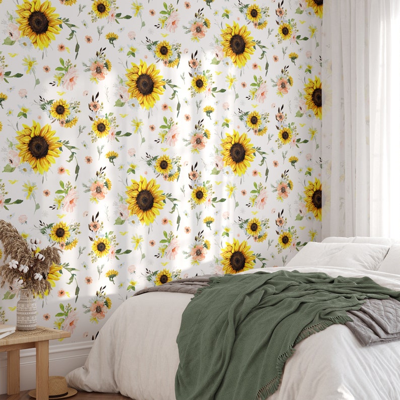 May include: Wallpaper featuring a repeating pattern of bright yellow sunflowers, soft pink flowers, and green foliage on a white background. The wallpaper is applied to a wall behind a bed with white bedding and a dark green throw blanket.