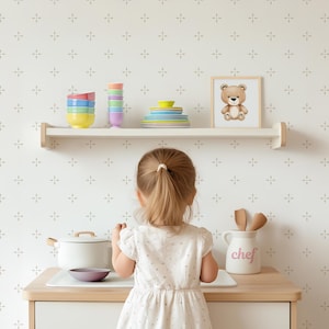 May include: A young child stands in front of a white countertop with a white pot, a pink bowl, and a white ceramic jar with the word "chef" on it. The countertop is in front of a white wall with a repeating pattern of small gray crosses. A shelf above the countertop holds a stack of colorful bowls and a stack of white plates.