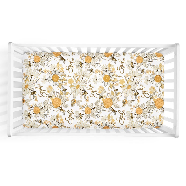 May include: A white crib sheet featuring a repeating pattern of yellow and white daisies, brown line-art leaves, and small bees. The name Sophie is printed in a brown cursive script throughout the design, set within a white wooden crib frame.