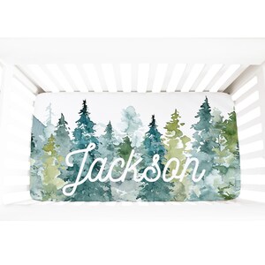 May include: A white crib sheet with a watercolor design of a forest with green and blue trees. The name "Jackson" is written in white cursive script on the sheet.