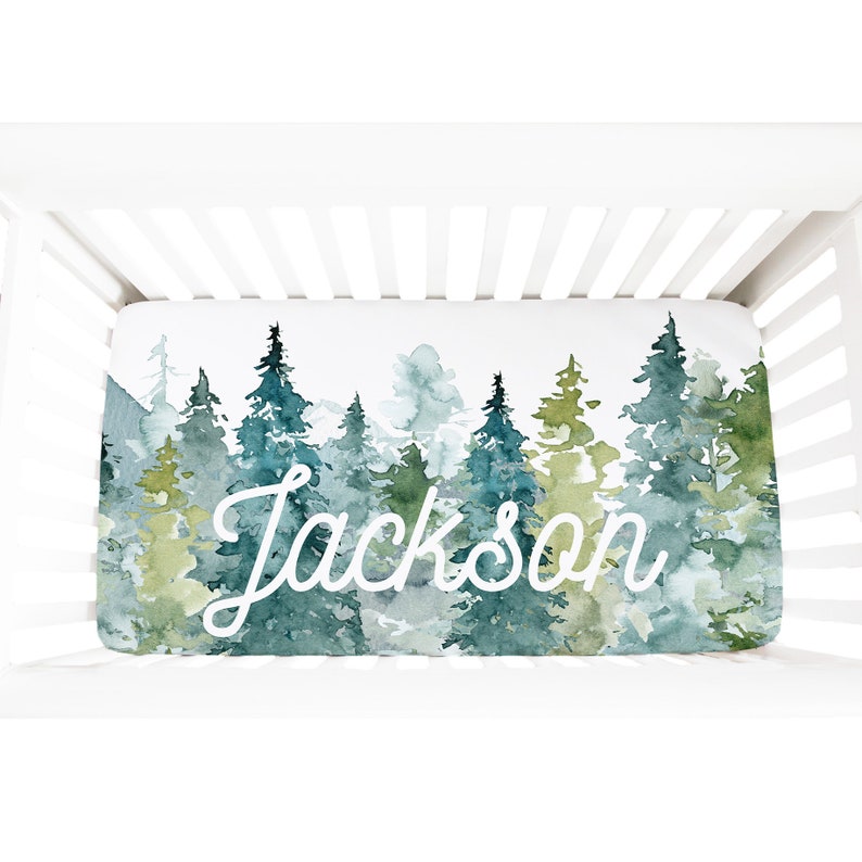 May include: A white crib sheet with a watercolor design of a forest with green and blue trees. The name "Jackson" is written in white cursive script on the sheet.