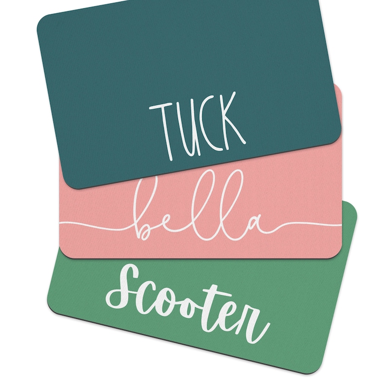 May include: Three rectangular name tags with white text on a green and pink background. The top tag is teal green with the name "Tuck" written in white. The middle tag is pink with the name "bella" written in white. The bottom tag is a darker green with the name "Scooter" written in white.