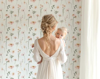 May include: A woman in a white dress holds a baby in her arms. The woman and baby are standing in front of a wall covered in a floral wallpaper with a repeating pattern of pink flowers and green leaves.