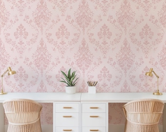 May include: A workspace featuring a long white desk with two white three-drawer cabinets, two light-colored woven rattan chairs, two gold desk lamps, a small potted green plant, and a pencil holder against a light pink damask patterned wallpaper.