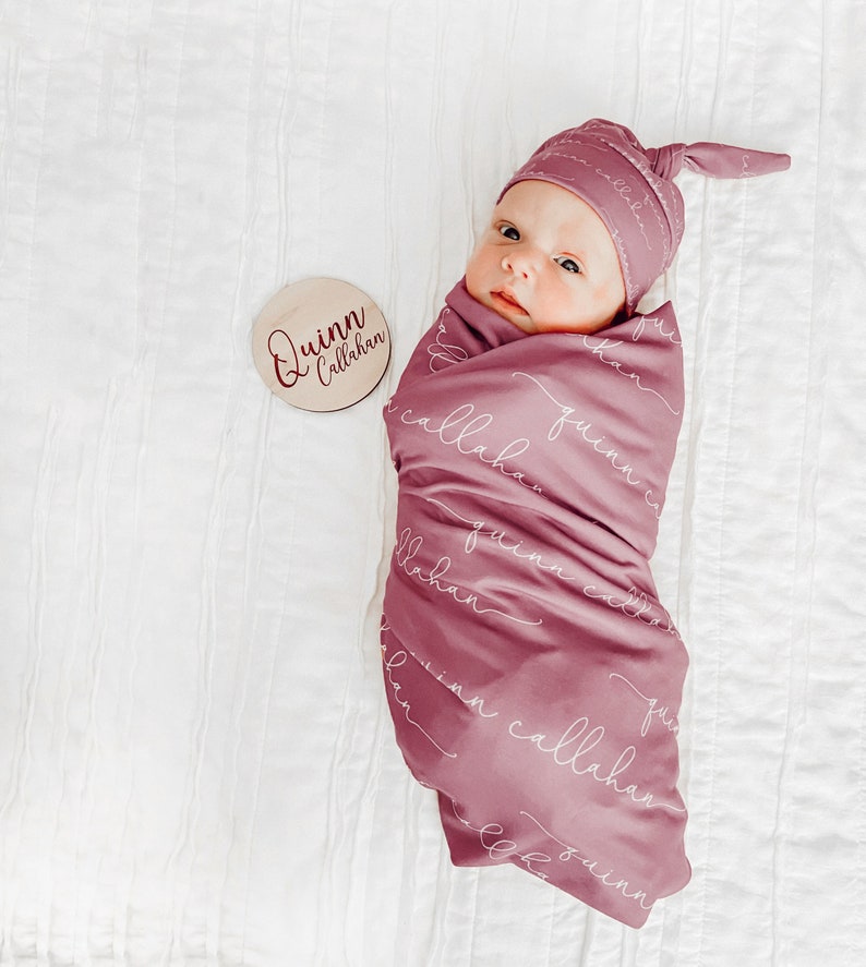 May include: A newborn baby swaddled in a pink muslin swaddle blanket with the name "Quinn Callahan" printed on it. The baby is wearing a pink knotted hat. A wooden circle with the name "Quinn Callahan" is in the background.
