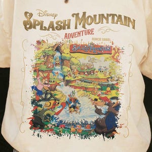 May include: Cream-colored t-shirt featuring a colorful graphic of Disney's Splash Mountain Adventure. The design includes various cartoon characters and the text "Disney Splash Mountain Adventure" in gold lettering. The shirt has short sleeves.