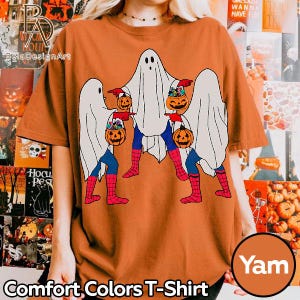 May include: An orange t-shirt featuring a graphic of three ghosts wearing white sheets over blue and red spider-web patterned leggings. Each ghost is holding an orange jack-o'-lantern bucket filled with colorful candy treats.