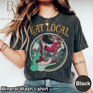 Puede incluir: Una camiseta negra con lavado mineral que presenta un gráfico circular con el texto EAT LOCAL. La ilustración muestra a un pirata de dibujos animados con abrigo rojo huyendo de un gran cocodrilo verde con la boca abierta, sobre un fondo tenue.