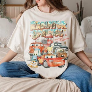 May include: A cream-colored short-sleeve t-shirt featuring a colorful graphic print of characters from the movie Cars. The text Radiator Springs and Lightning McQueen is printed above the characters, which include a red race car and a gold trophy.