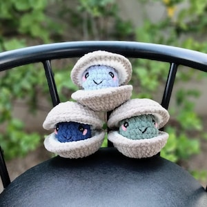 May include: Three handmade crochet clam shells, each with a smiling face peeking out. The clamshells are a light beige colour, and the creatures inside are blue, navy, and green. The background is a blurred green.