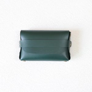 May include: A dark green leather wallet with a strap closure. The wallet is rectangular with rounded edges and brass rivets. The strap wraps around the wallet and fastens with a brass snap. The leather has a smooth, matte finish.