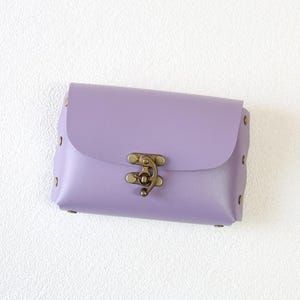 May include: A small, rectangular lavender-colored purse with a flap closure. The purse features a decorative antique brass clasp and is secured with small rivets along the edges. The purse is made of a smooth, solid material.