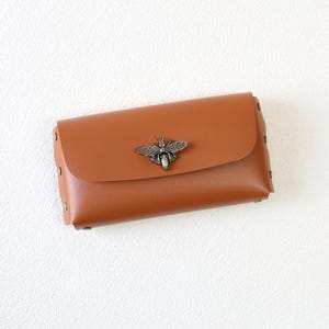 May include: A brown leather clutch with a flap closure and a decorative metal bee emblem. The rectangular bag has a series of small rivets along the edges. The clutch is a warm, neutral color.