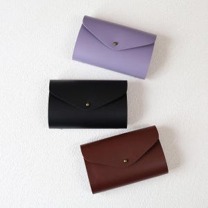 以下が含まれることがあります： ラベンダー、黒、茶色の3色の革製封筒型財布。各財布は、小さな丸い真鍮色のボタンで留められたフラップクロージャーを備えています。財布は白いテクスチャのある表面に配置されています。