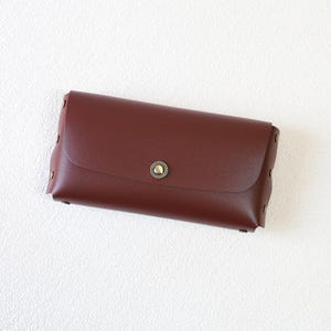May include: A burgundy-colored, rectangular clutch with a rounded flap and a brass-colored button closure. The clutch is adorned with small, brass-colored rivets along the edges. The design is simple and elegant, suitable for various occasions.