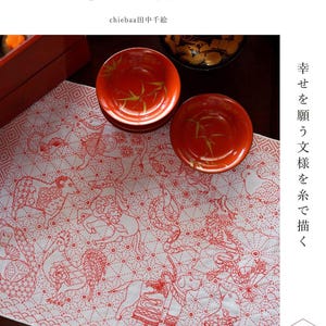 May include: Two red ceramic bowls with gold bamboo leaf designs sit atop a white cloth with red embroidered animal patterns. The cloth features various animals and geometric designs. Japanese text is visible.