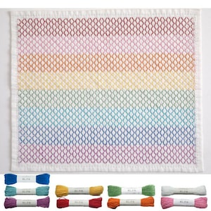 May include: A white, square, embroidered cloth with a rainbow pattern. The design features a grid of diamond shapes in red, orange, yellow, green, blue, and purple. Below are spools of thread in matching colors.
