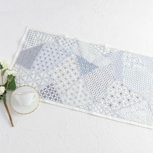 May include: A white and blue patterned table runner with a patchwork design. The runner features various geometric patterns in shades of blue and white, with a fringed edge. A white teacup and saucer are on the left.