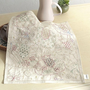 Può includere: Tovaglietta in lino color crema con ricami colorati e intricati. Il design presenta motivi geometrici e floreali in filo rosso, verde, blu e oro. I bordi sono ben cuciti e una piccola etichetta è visibile in basso a destra.