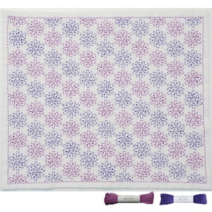 Puede incluir: Textil cuadrado blanco con un patrón repetido de bordados florales morados y rosas. El bordado tiene un diseño de estrella. Dos carretes de hilo, uno morado y otro violeta, están en la parte inferior.