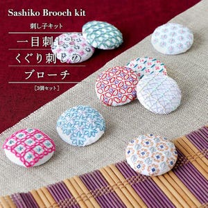 Op de afbeelding: Een verzameling geborduurde broches in verschillende kleuren en patronen. De ronde broches hebben gedetailleerde steken in roze, blauw, rood en wit. De tekst "Sashiko Brooch kit" is zichtbaar.