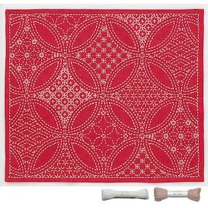 Puede incluir: Un textil rectangular rojo con un patrón cosido blanco de círculos superpuestos y diseños florales. El textil es probablemente una tela para manualidades o decoración del hogar. Dos carretes de hilo en la parte inferior.