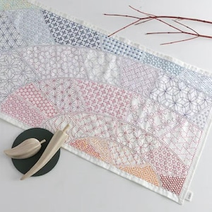 Op de afbeelding: Een witte en veelkleurige gewatteerde tafelloper met ingewikkelde gestikte patronen in een waaierachtig ontwerp. De loper heeft tinten blauw, roze, oranje en wit. Een houten blad en een pepervormig object van hout liggen op een zwarte cirkelvormige plaat.