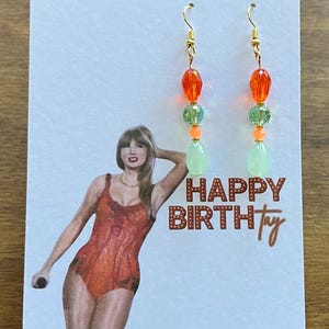 May include: A pair of dangle earrings with gold-colored hooks. Each earring features orange, green, and light green beads. The earrings are displayed on a card with a woman in an orange swimsuit and the text "HAPPY BIRTHtay".