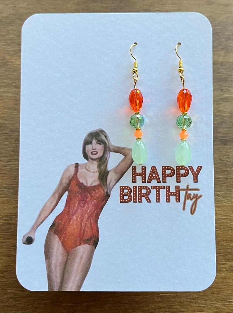 May include: A pair of dangle earrings with gold-colored hooks. Each earring features orange, green, and light green beads. The earrings are displayed on a card with a woman in an orange swimsuit and the text "HAPPY BIRTHtay".
