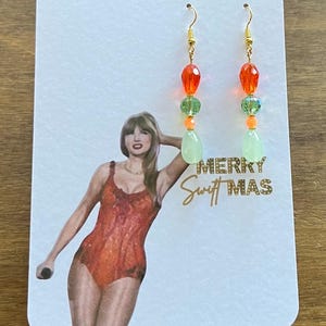 May include: A pair of dangle earrings with gold-colored hooks. Each earring features orange, green, and light green beads. The earrings are displayed on a white card with a photo of a person and the text "MERRY Swift MAS".