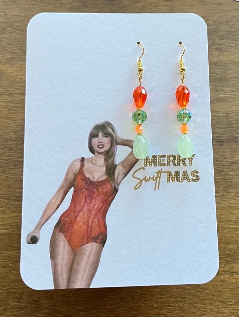 May include: A pair of dangle earrings with gold-colored hooks. Each earring features orange, green, and light green beads. The earrings are displayed on a white card with a photo of a person and the text "MERRY Swift MAS".