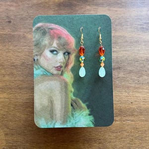 May include: Dangling earrings with gold-colored hooks. Each earring features a series of orange, green, and clear beads, culminating in a teardrop-shaped, pale green bead. The earrings are displayed on a card with a portrait of a woman.