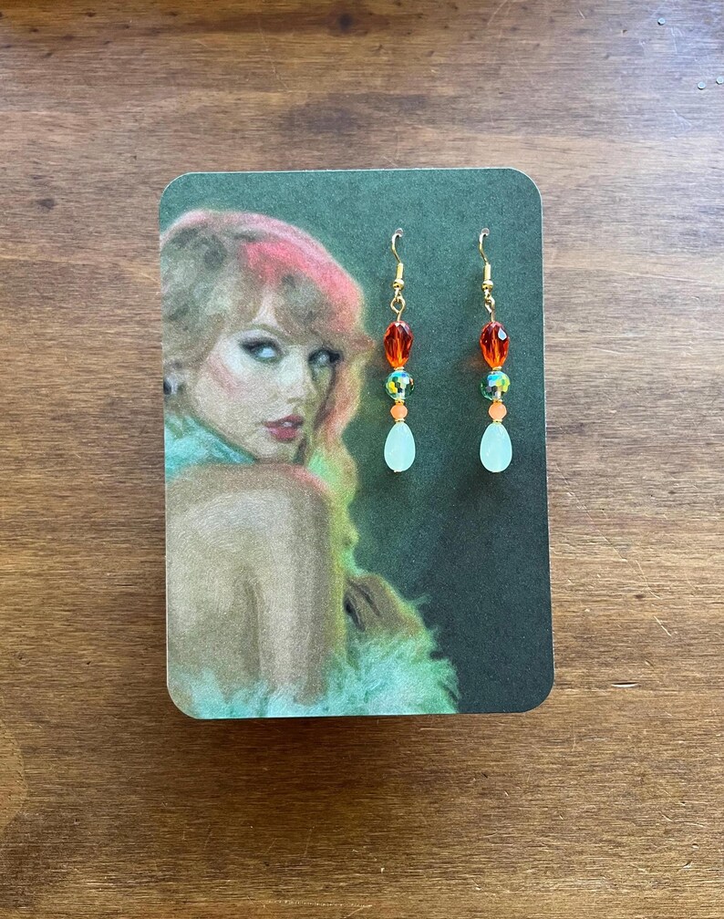 May include: Dangling earrings with gold-colored hooks. Each earring features a series of orange, green, and clear beads, culminating in a teardrop-shaped, pale green bead. The earrings are displayed on a card with a portrait of a woman.