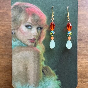 May include: A pair of dangle earrings with gold-colored hooks. Each earring features a red, orange, and iridescent bead arrangement, culminating in a teardrop-shaped, pale green bead. The earrings are displayed on a card with a portrait.