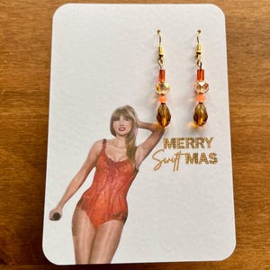 May include: A pair of dangle earrings with gold-colored hooks and orange and amber-colored beads. The earrings are displayed on a white card with a picture of a person in an orange outfit and the text "MERRY Swift MAS".