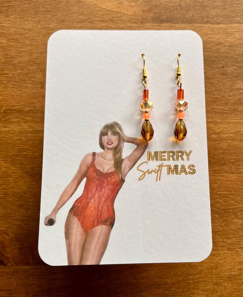 May include: A pair of dangle earrings with gold-colored hooks and orange and amber-colored beads. The earrings are displayed on a white card with a picture of a person in an orange outfit and the text "MERRY Swift MAS".